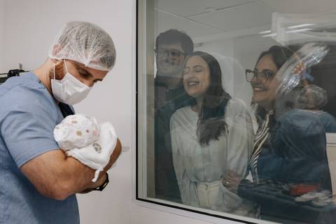 Pai apresentando o bebê para os familiares através do vidro.'