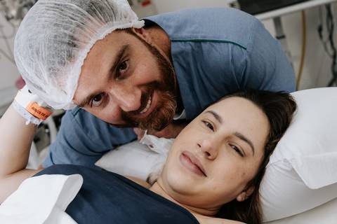 Pai e mãe deitados sorrindo para a câmera após o parto.'