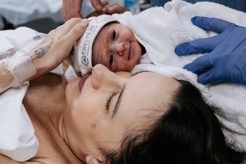 Bebê no peito da mãe olhando para a câmera com apoio do pai.'