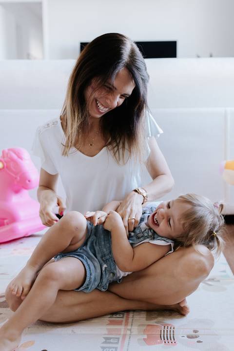 Mãe fazendo cosquinha na filha deitada no chão, com selo no canto superior.'