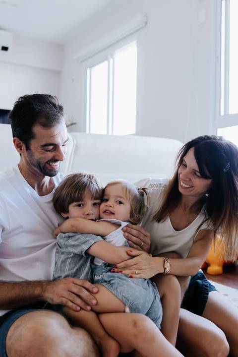 Pais abraçados aos dois filhos sorrindo no chão da sala.'