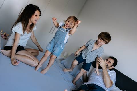 Crianças pulando na cama com os pais observando ao fundo.'