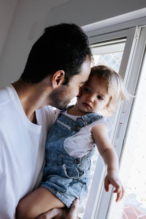Pai segurando a filha no colo perto da janela.'