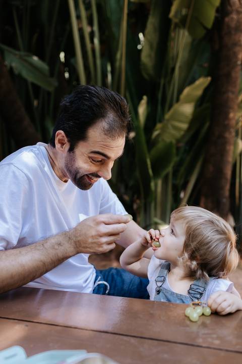 Pai dando um frutinha na boca da filha no parque.'