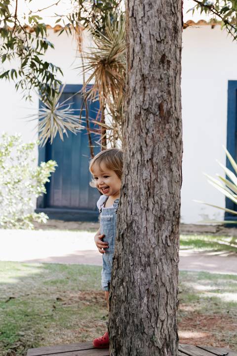 Menina espiando sorridente atrás do tronco da árvore.'