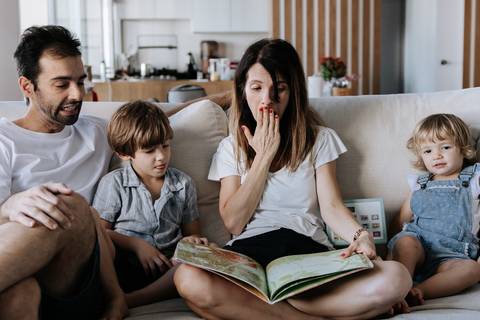 Mãe lendo um livro infantil para os filhos e o pai no sofá.'