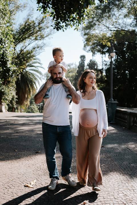 Pai com o filho nos ombros caminhando ao lado da gestante no parque.'