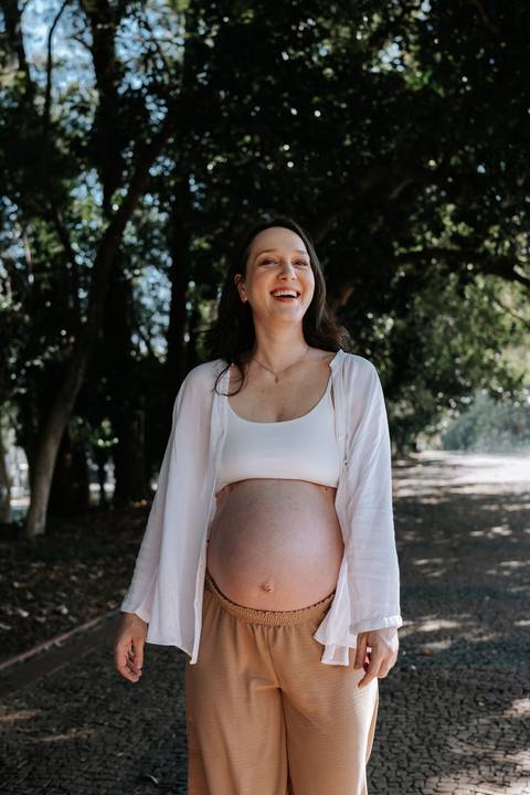 Retrato em plano médio da gestante sorrindo em meio à natureza.'