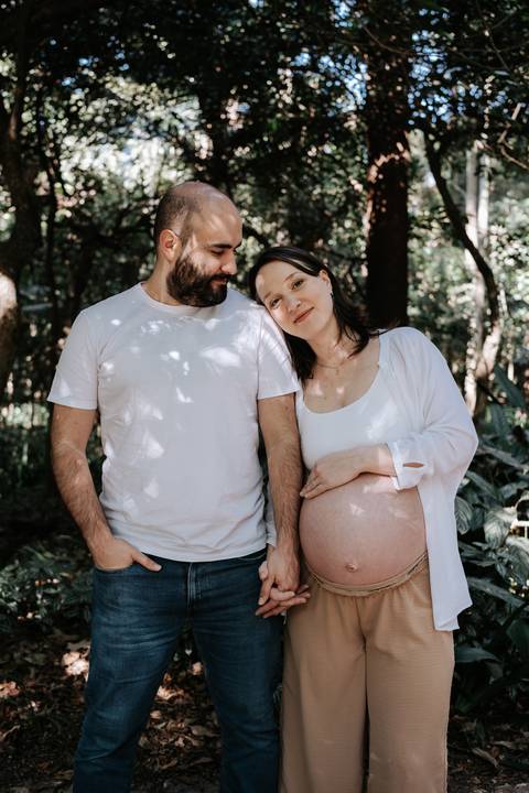 Casal de branco posando lado a lado no bosque, com a mãe segurando a barriga.'
