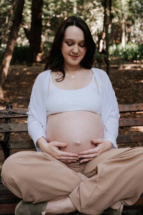 Gestante sentada no banco do parque com as mãos na barriga.'