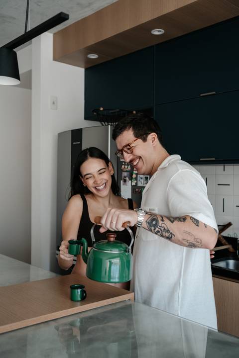 Casal sorrindo e preparando café na cozinha.'