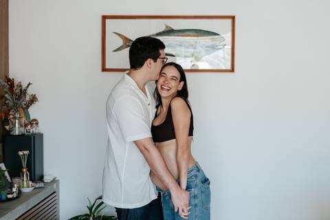 Casal abraçado e sorridente posando em pé na sala.'