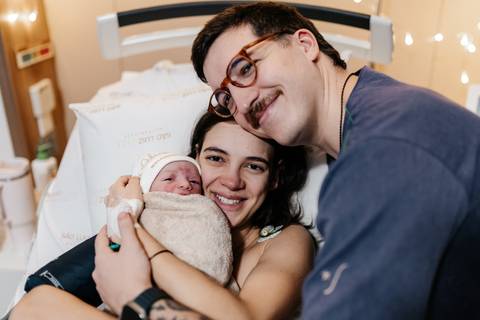 Retrato do casal sorrindo abraçados com o bebê enrolado na manta.'