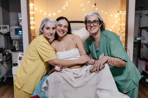 Mãe sorridente abraçada com a equipe médica na sala de parto.'