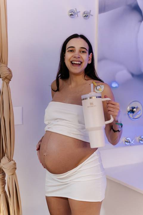 Gestante sorrindo com copo térmico na sala de parto.'
