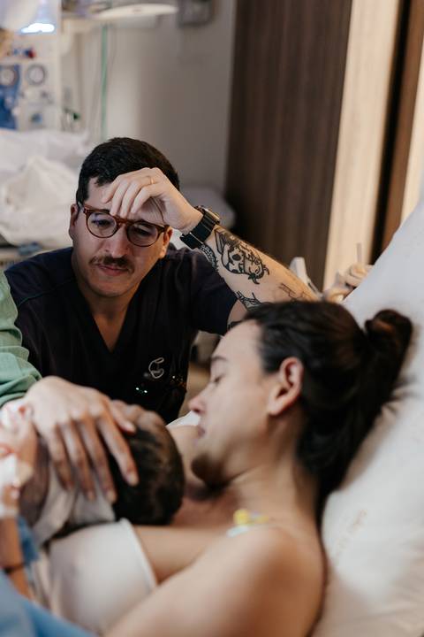 Pai emocionado com a mão no rosto ao lado da mãe e do bebê recém-nascido.'