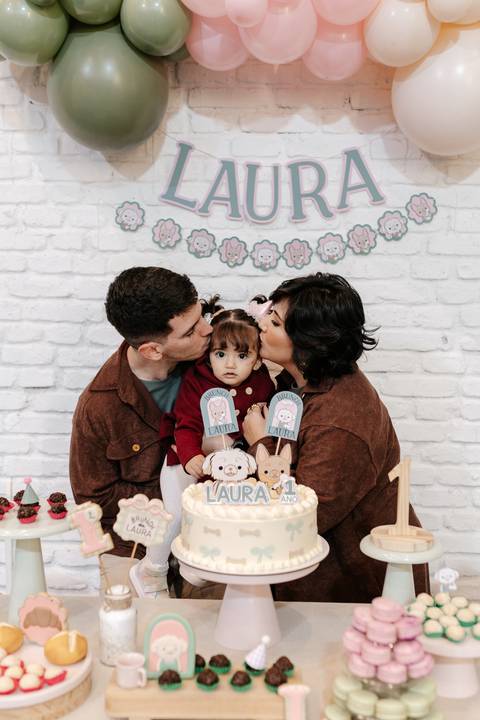 Pais beijando as bochechas da aniversariante atrás do bolo.'