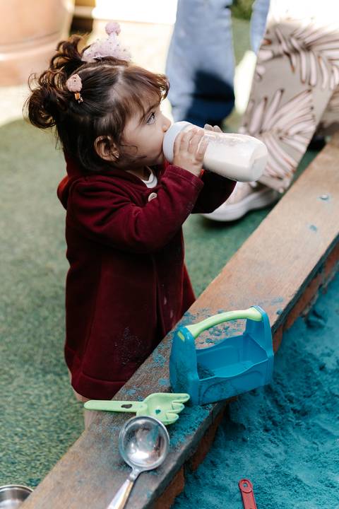 Aniversariante tomando mamadeira em pé perto dos brinquedos.'