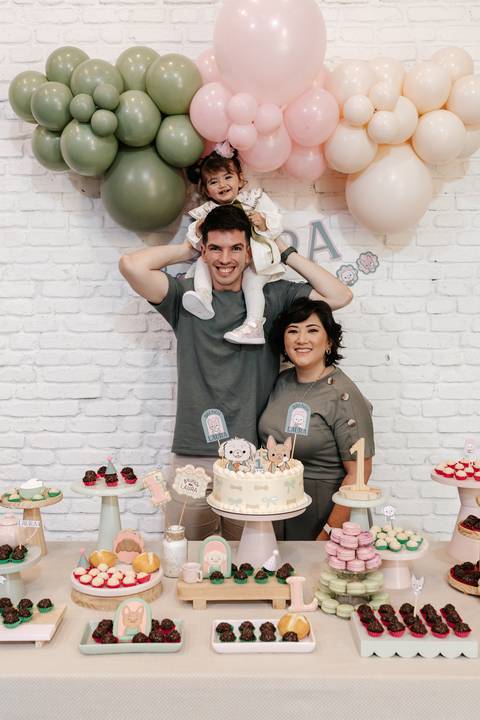 Pai com a aniversariante nos ombros e a mãe ao lado na mesa do bolo.'