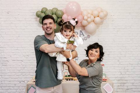 Pais erguendo a aniversariante em vestidinho claro em frente ao painel.'