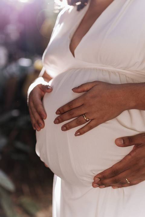 Close-up das mãos do casal entrelaçadas sobre a barriga de grávida.'