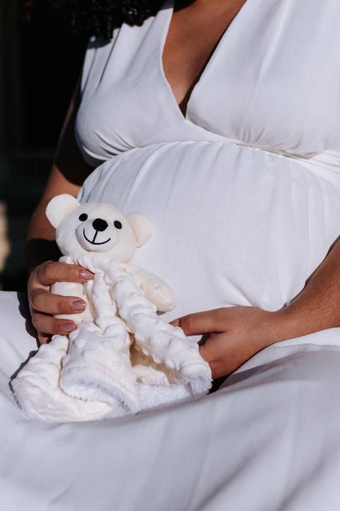Detalhe das mãos da gestante segurando um ursinho de pelúcia na barriga.'
