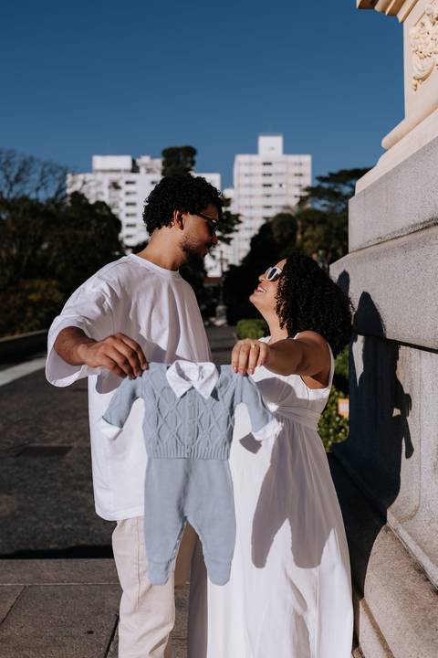 Casal segurando um macacãozinho azul de bebê em frente à arquitetura.'