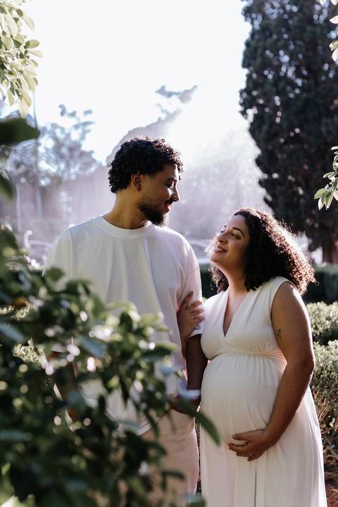 Casal sorrindo entre as folhagens do parque com iluminação natural.'
