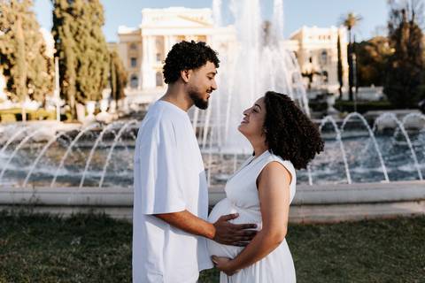 Casal de frente para a fonte d'água com o pai segurando a barriga da mãe.'