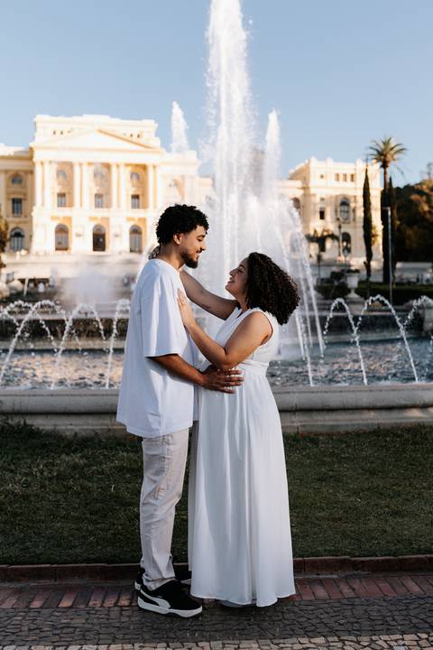 Casal abraçado sorrindo com os chafarizes do parque ao fundo.'