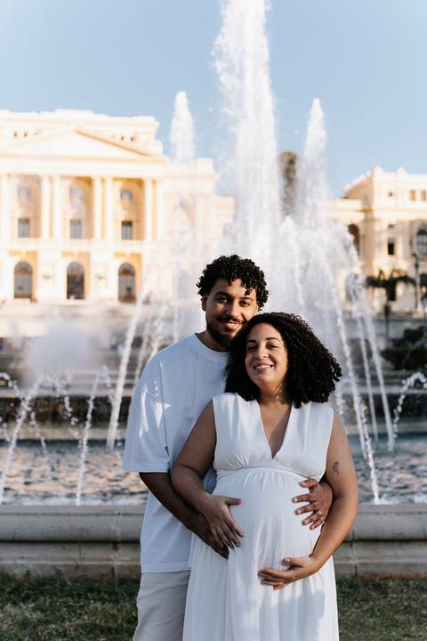 Casal sorridente posando juntos em frente à fonte de água do parque.'