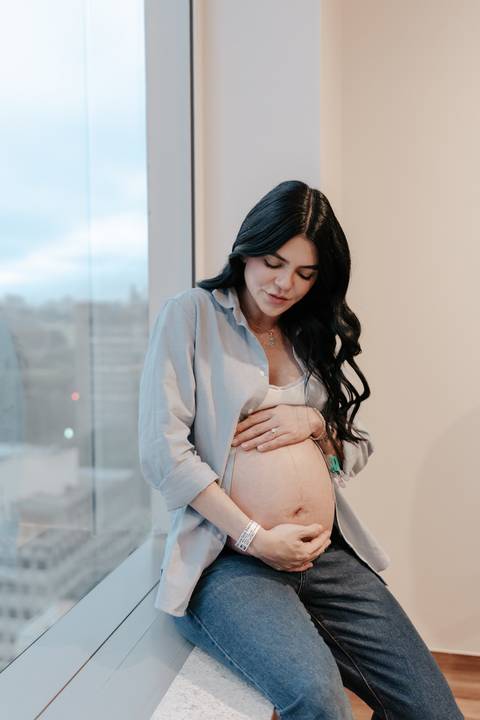 Gestante de camisa jeans aberta olhando carinhosamente para a barriga.'