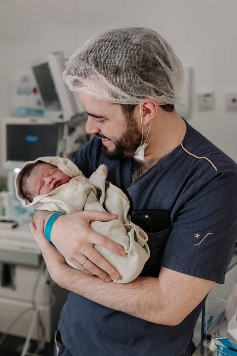 Pai sorrindo orgulhoso segurando o bebê envolto em mantas no colo.'