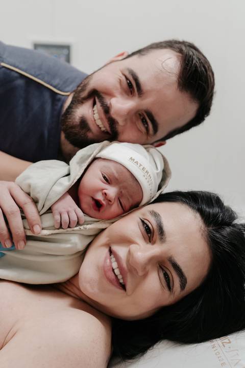Retrato do casal sorrindo juntinho com o bebê no colo.'
