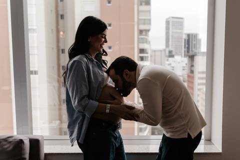 Pai beijando a barriga da gestante em frente à janela com vista urbana.'