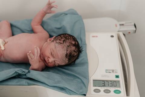 Bebê recém-nascido sendo pesado na balança pediátrica da sala de parto.'