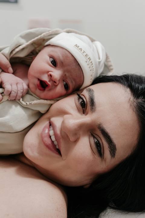 Mãe sorrindo radiante deitada com o bebê acordado ao seu lado.'