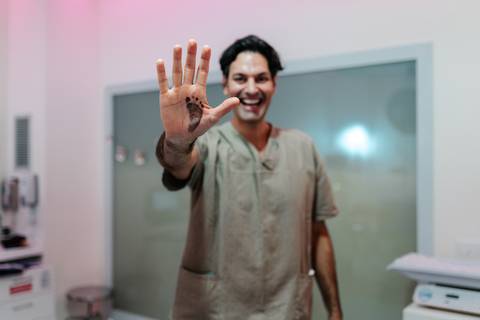 Pai sorridente fazendo o sinal do número 5 com a mão na sala de parto.'