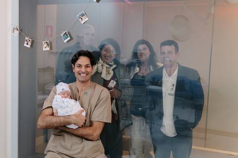 Pai segurando o recém-nascido no colo tirando foto com a família através do vidro. '