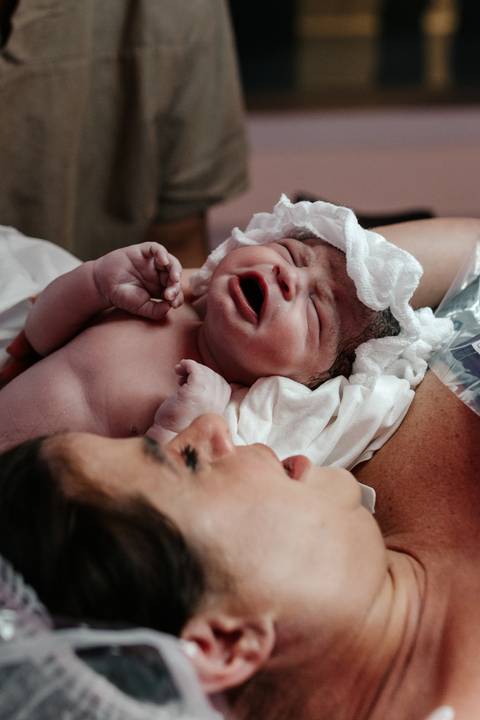 Mãe olhando com carinho para o bebê recém-nascido chorando no seu peito.'