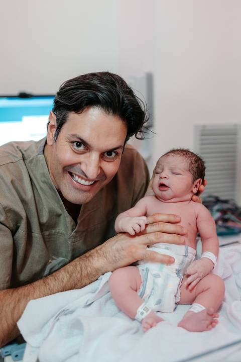 Pai sorrindo orgulhoso segurando o bebê no colo após as avaliações.'
