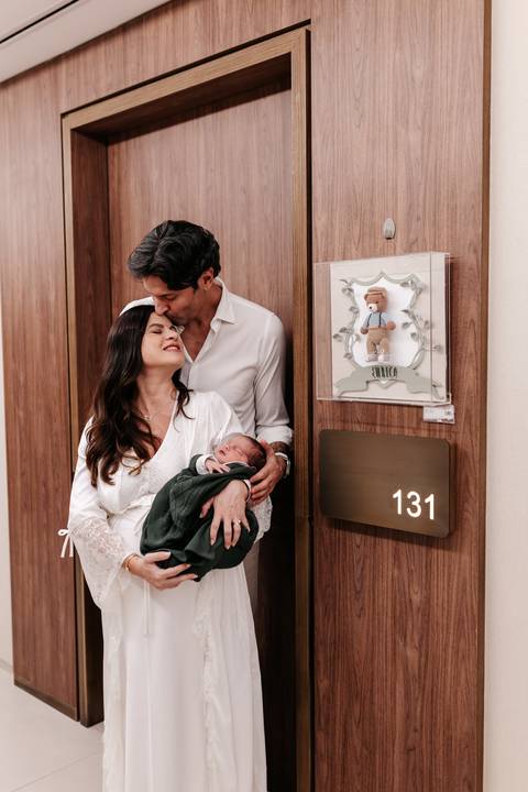 Pai beijando a cabeça da mãe enquanto ela segura o bebê na porta do quarto 131.'