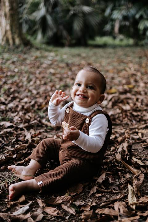 Rafael sorrindo sentado entre as folhas durante ensaio fotográfico de 1 ano'