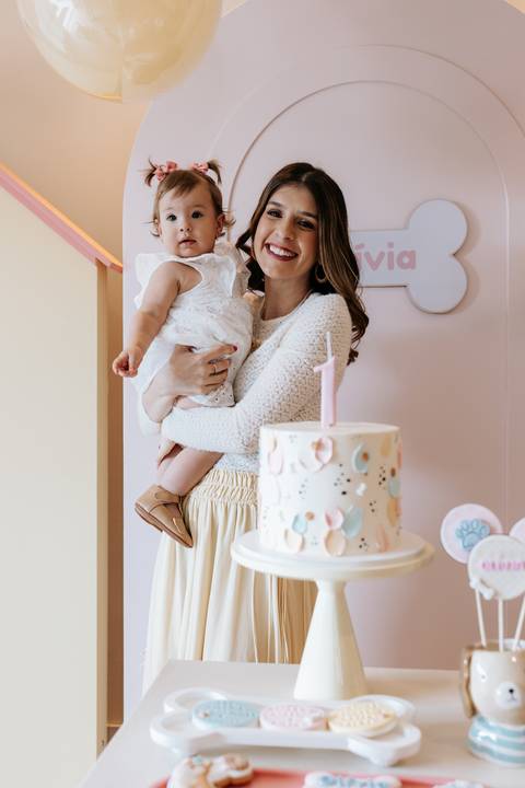 Olívia no colo da mãe durante sua festa de 1 ano, ao lado do bolo decorado com o tema cachorrinho.'