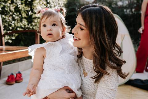 Olívia no colo da mãe durante sua festa de 1 ano, sorrindo em um espaço externo decorado.'