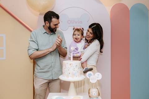 Olívia sorrindo entre os pais diante do bolo durante a festa de 1 ano.'
