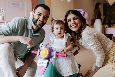Olívia posa com os pais enquanto brinca em um brinquedo infantil durante a comemoração de seu primeiro aniversário.'