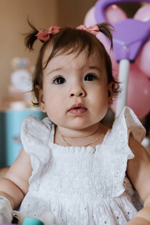 Close do rosto de Olívia, de vestido branco, olhando atentamente para a câmera durante sua festa de 1 ano.'