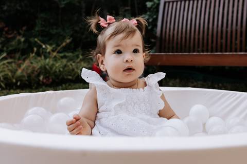 lívia sentada em uma piscina de bolinhas brancas durante a comemoração de seu primeiro aniversário.'