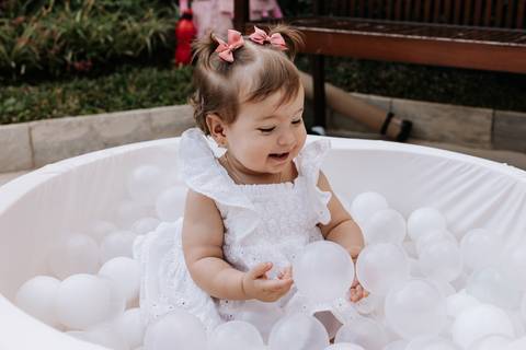 Olívia brinca em uma piscina de bolinhas brancas durante sua festa de 1 ano, usando vestido branco e laços rosas no cabelo.'